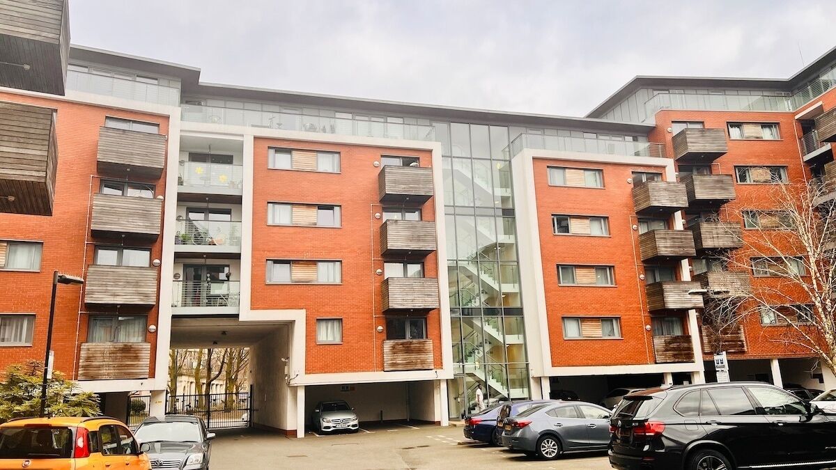 A modern apartment building with multiple balconies, a central glass elevator shaft, and a parking area with several cars.