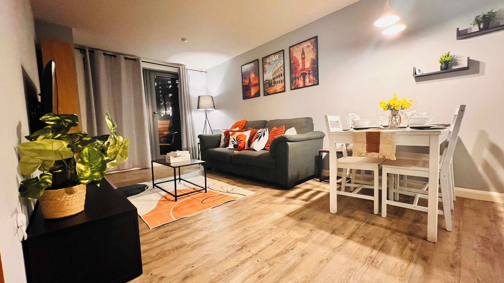 A modern living room with a sofa, dining table, and chairs, a potted plant, and a cityscape poster on the wall.