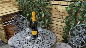 Een ronde metalen tafel met ingewikkelde bloemdecoraties, twee stoelen met beklede zittingen, een fles champagne en twee glazen.