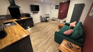 Een moderne keuken met een ingebouwde oven, een eethoek met een kleine tafel en stoelen, en een zithoek met een groene bank en een houten salontafel.
