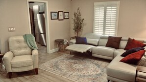 A living room with a beige sofa, a white armchair, a coffee table, and a patterned rug. There is a potted plant and a small side table. - Midtown Cottage, unique stone patio, family-dog friendly newly remodeled. (Tulsa)