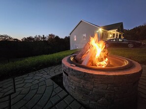 A stone fire pit with a burning fire, surrounded by a paved walkway, in front of a house with a lit exterior. - Hunter House (Livingston Manor)