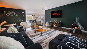 Moderni olohuone, jossa on musta sohva, sohvapöytä ja takka. Televisio on kiinnitetty seinään, ja siinä on kyltti, jossa lukee ”NETFLIX”.
