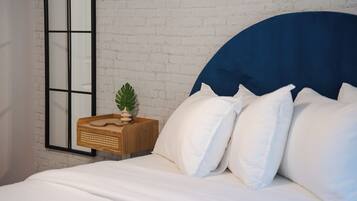 Une chambre avec un mur en briques blanches, une grande fenêtre à cadre noir, une table de chevet en bois ornée d’une plante, et un lit avec une literie blanche et une tête de lit bleue.