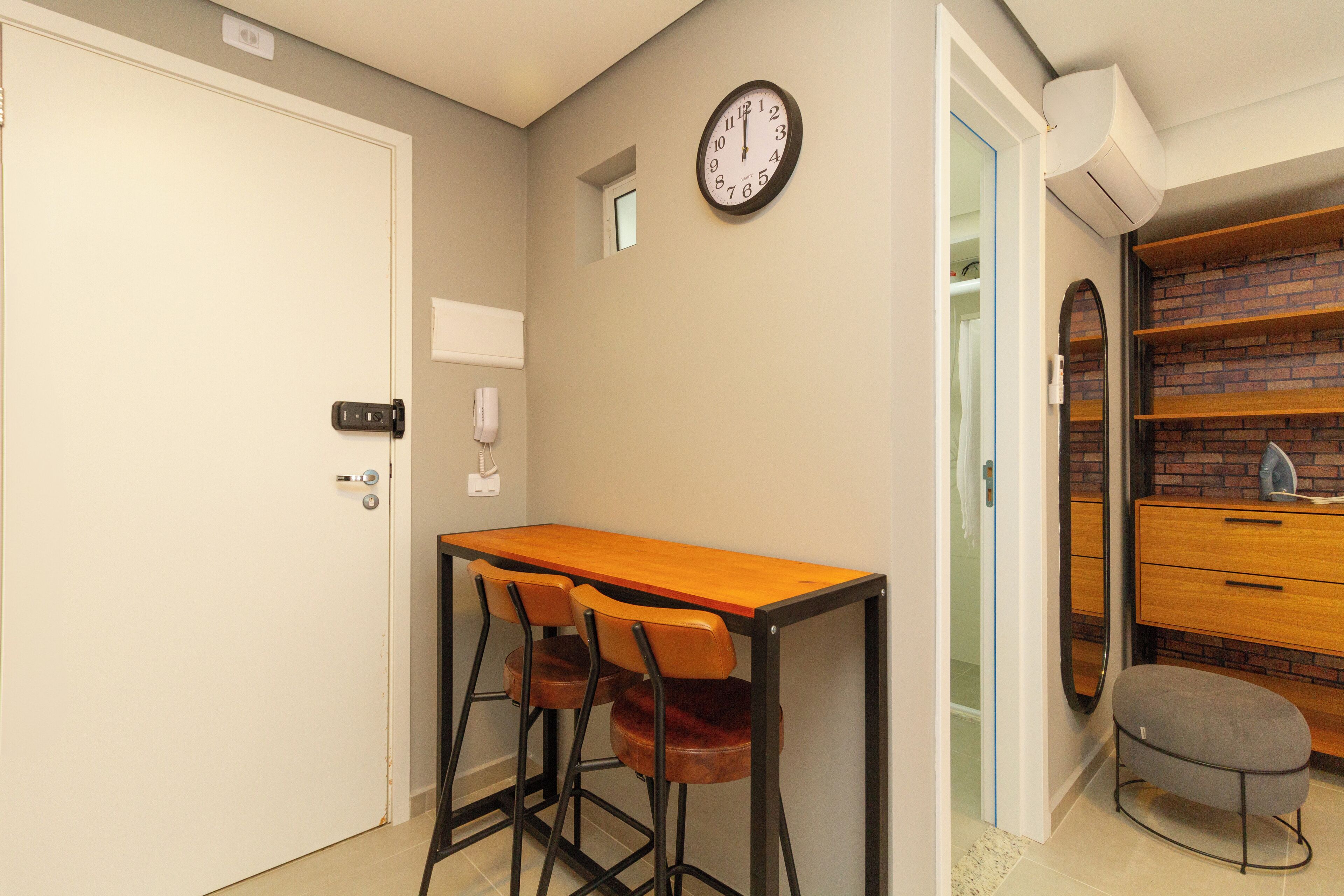 A small room with a wooden table and two chairs, a clock on the wall, and a brick-patterned shelf.