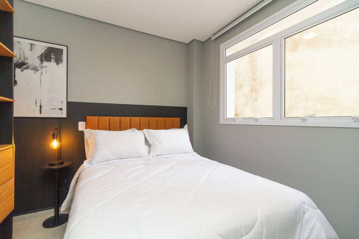 Un lit bien fait, avec du linge de lit blanc, dans une pièce dont le mur est gris et qui possède une fenêtre.
