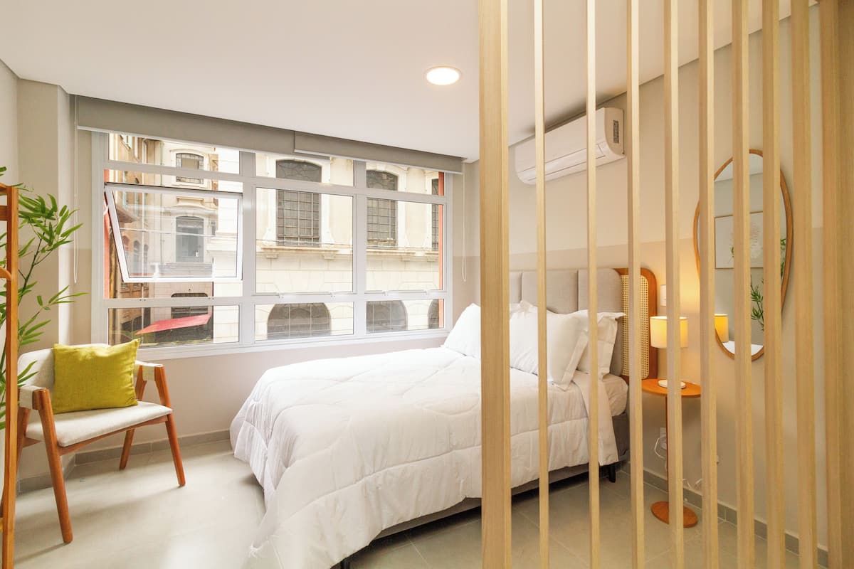 Une chambre avec une grande fenêtre, un lit, une chaise et une table de chevet.