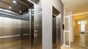 Un ascensore moderno con pareti metalliche e un pannello di controllo, che conduce a un corridoio con una porta in vetro e pavimento piastrellato.
