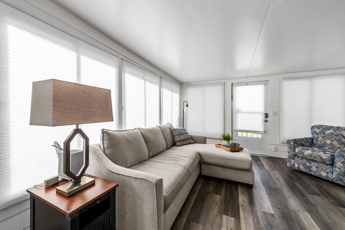 A modern living room with a beige sectional sofa, a patterned armchair, a wooden side table with a lamp, and a small plant on a tray.