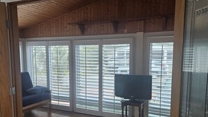 A room with wooden floors, a television on a stand, and a blue chair near a large window with blinds. - Oceanside Village Beautiful 3BR 3BA - Golf Cart Included! (Surfside Beach)