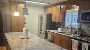 A modern kitchen with a granite countertop, stainless steel appliances, wooden cabinets, and pendant lighting. - Oceanside Village Beautiful 3BR 3BA - Golf Cart Included! (Surfside Beach)