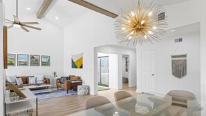 A spacious living room with a modern chandelier, a dining area with glass tables, and a cozy seating area with a brown sofa and patterned cushions. - The Palmer! Monterey Country Club, Hole 6, Resort Amenities! Golf cart included! (Palm Desert)