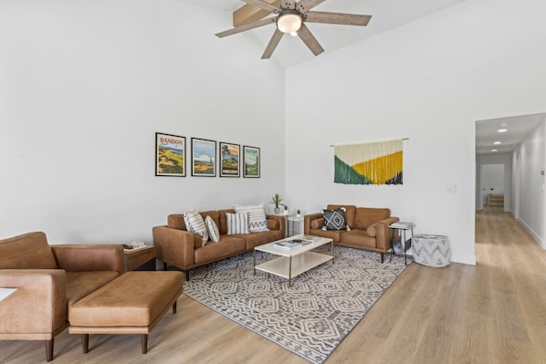 A spacious living room with a brown leather sofa, a coffee table, and a patterned rug. There are framed pictures on the wall and a ceiling fan.