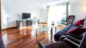 Ett modernt vardagsrum med en soffa, ett soffbord i glas, en tv placerad på en tv-bänk och utsikt över utomhusmiljön genom stora fönster.