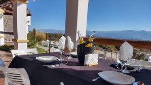 Uma área de refeições ao ar livre com uma mesa posta para dois, uma garrafa de vinho branco e vista para montanhas ao fundo.