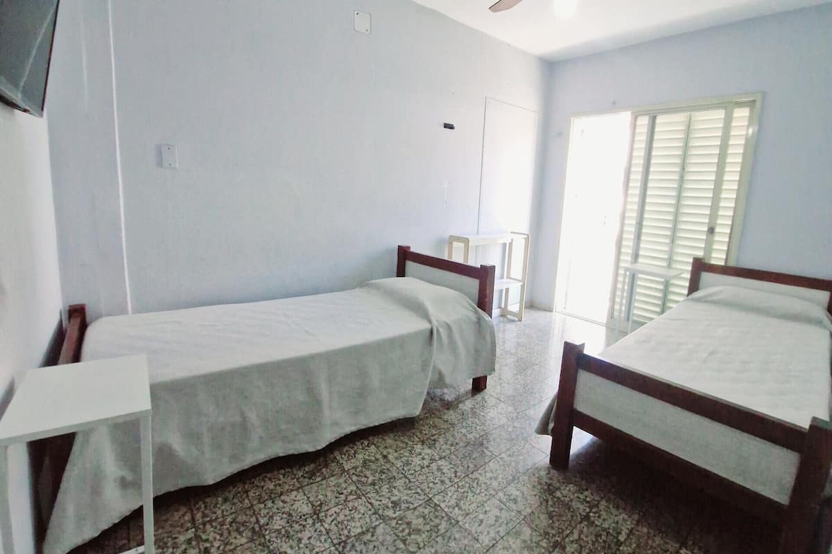 Una habitación con dos camas individuales, un escritorio, una silla, un ventilador de techo y una ventana con persianas.