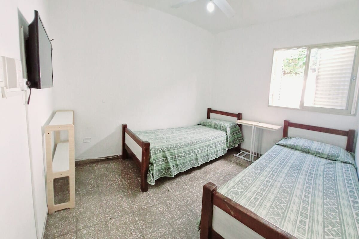 Una habitación pequeña y limpia con dos camas individuales, un ventilador de techo, una ventana con persianas y un televisor montado en la pared.