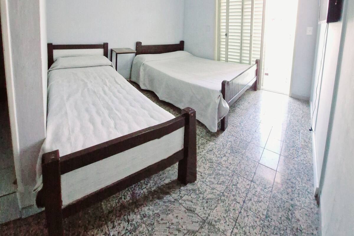 Una habitación con dos camas individuales, ventilador de techo, ventana con persianas y suelo de baldosas.