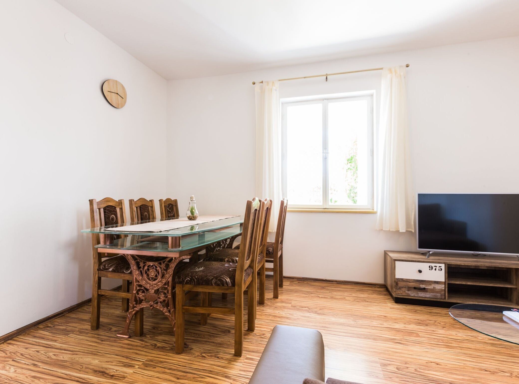 Un espacio habitable compacto con una mesa de comedor de vidrio rodeada de sillas de madera, un televisor sobre un mueble de madera, y una ventana con cortinas.