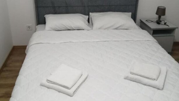 A neatly made bed with white linens and pillows, a bedside table with a lamp, and a small drawer unit.