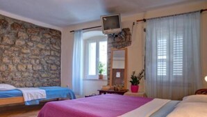 A bedroom with two beds, a stone wall, a television, and a window with curtains. - Quaint & Affordable Stay in Budva Traditional Stone House - Triple Room 5 (Budva)
