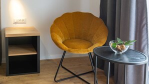 Une chaise de couleur moutarde avec un cadre en métal noir, une petite table en bois sur laquelle se trouve un bol en verre et quelques feuilles vertes, et une prise de courant murale.