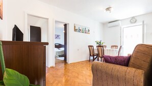 Una sala de estar con piso de madera, un sofá, una mesa de comedor con sillas y una cocina visible a través de un umbral abierto.