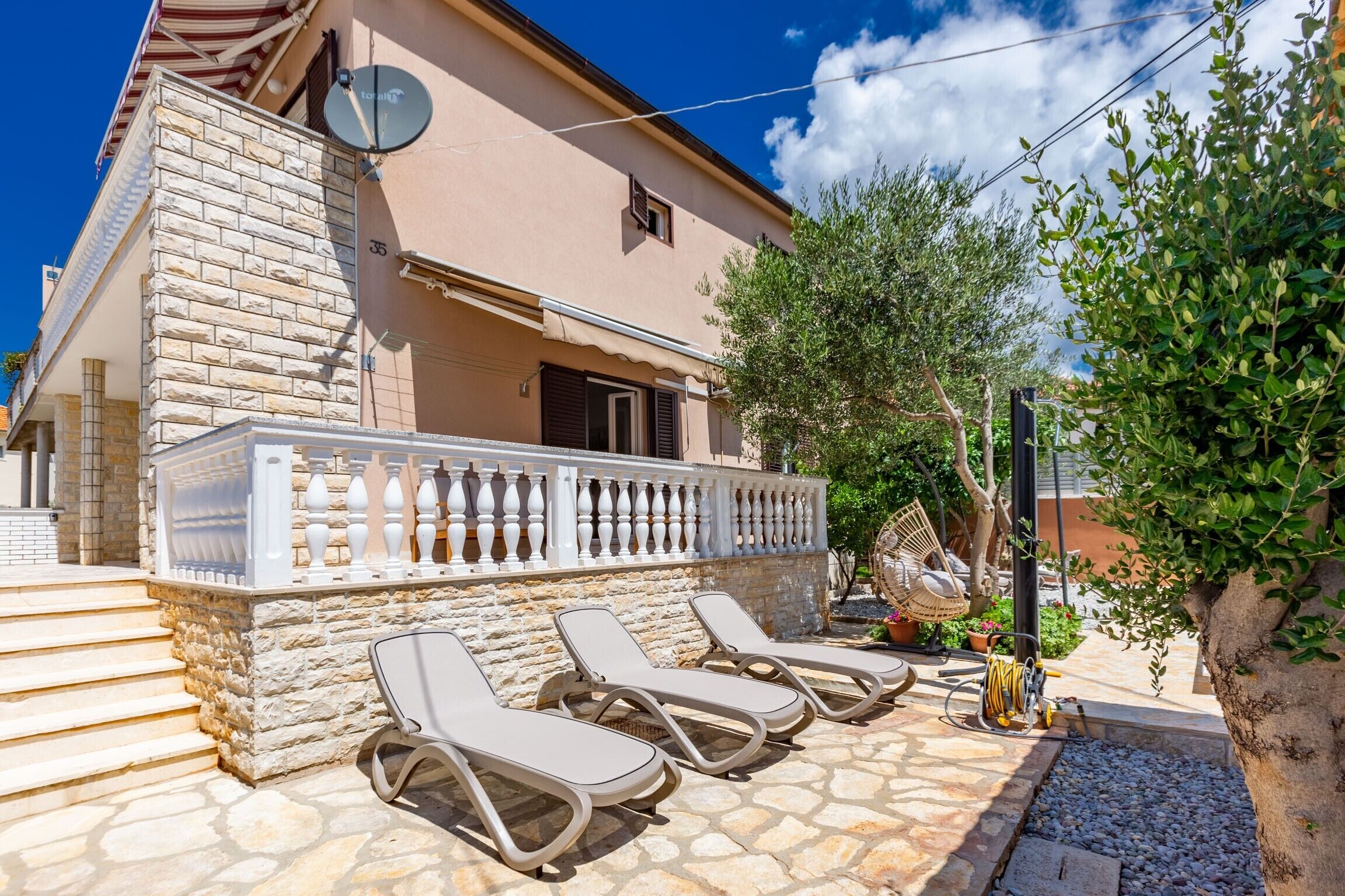 A building with a stone facade, a balcony with white balustrades, and three sun loungers on a stone patio.