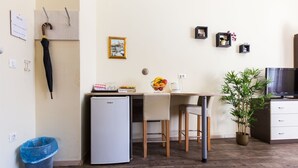 A compact kitchen with a refrigerator, a dining table with chairs, a small trash bin, and a potted plant.