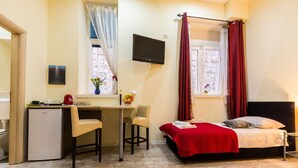 Habitación de hotel compacta con cama, escritorio, refrigerador pequeño y televisor montado en la pared.