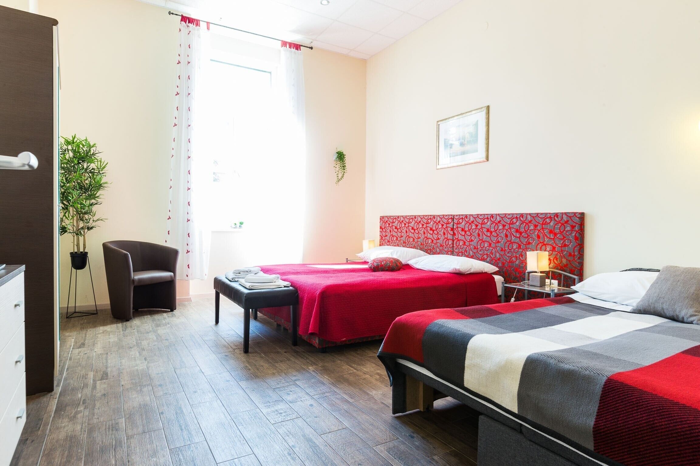 Une chambre d’hôtel avec deux lits, un bureau, une chaise, une plante en pot et une fenêtre avec des rideaux.