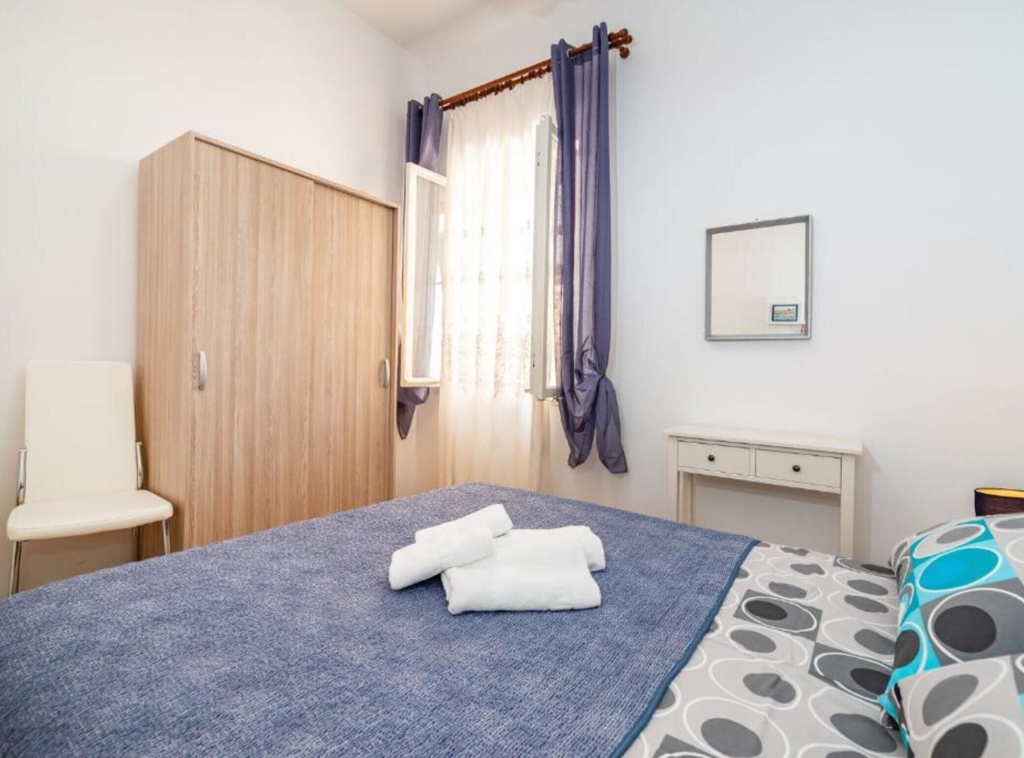 Un dormitorio con cama, una silla, armario, mesita de noche y ventana con cortinas.