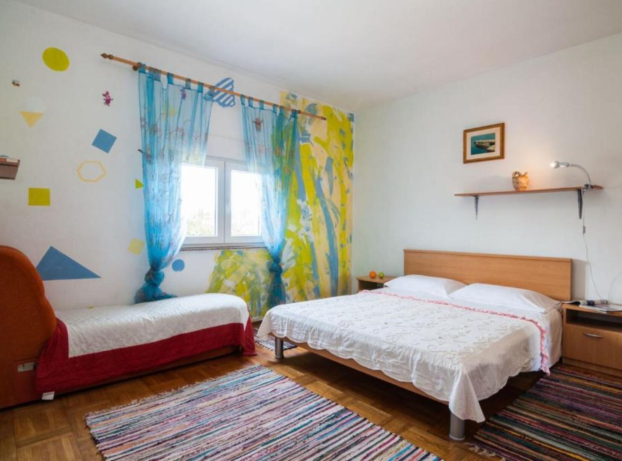 A bedroom with two single beds, a window with curtains, a bedside table with a lamp, and a small shelf with a picture.