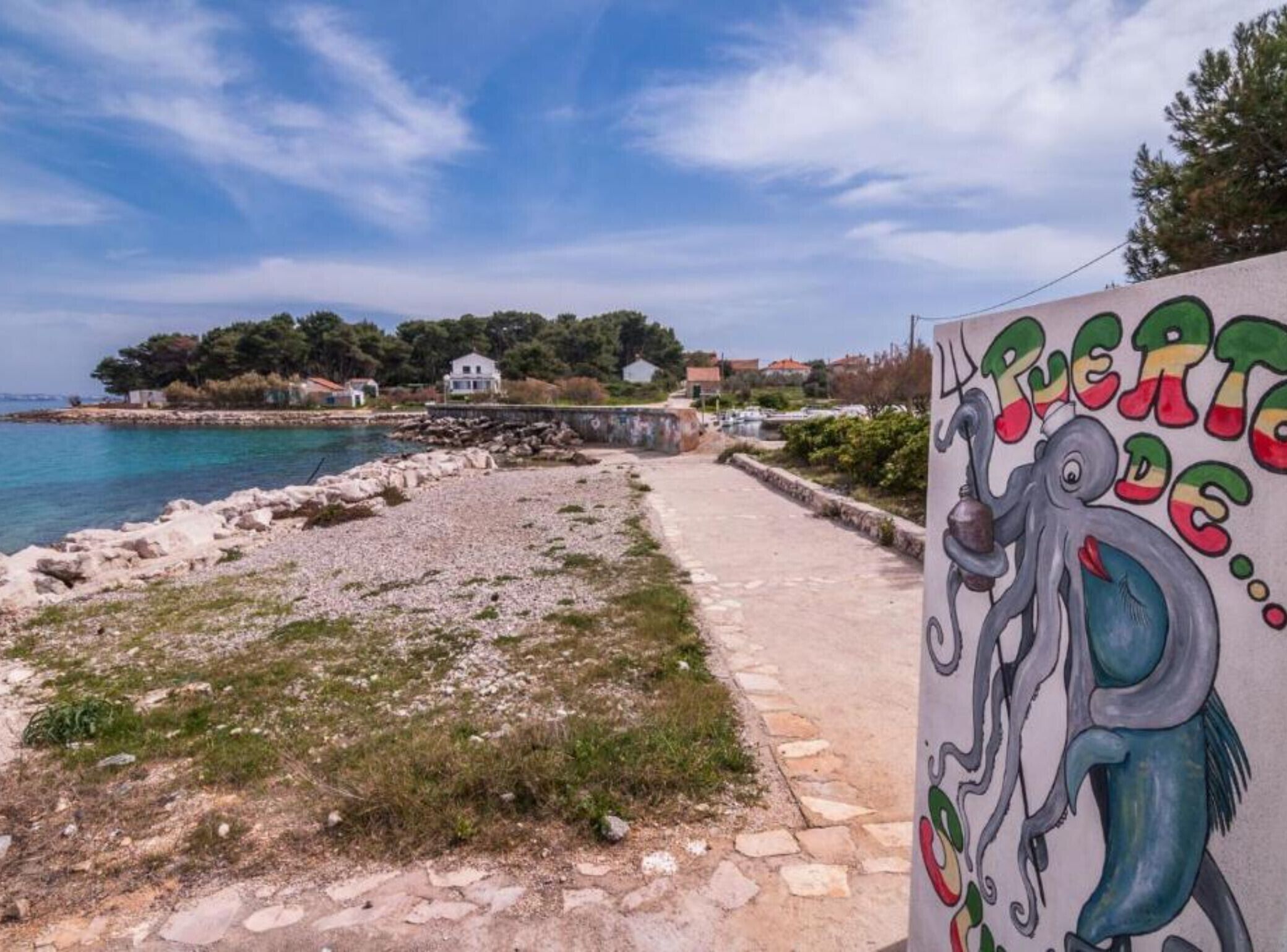 A coastal path with a rocky beach, a clear blue sky, and a colorful mural of an octopus.