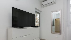 Een slaapkamer met een grote flatscreen televisie op een witte dressoir, een raam met uitzicht op bomen, en een spiegel aan de muur.