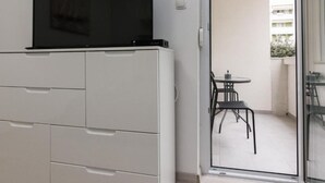 Een modern appartement met een flatscreen televisie op een witte kast, een balkon met een tafel en stoelen, en uitzicht op een gebouw op de achtergrond.