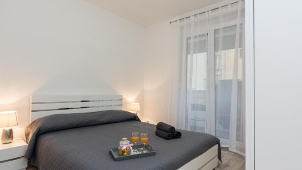 Een moderne hotelkamer met een groot bed, een nachtkastje met een lamp, en een dienblad met drankjes en een kleine fles.