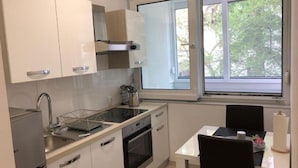Una cocina moderna con armarios blancos, un lavamanos de acero inoxidable y un horno. Hay una ventana que proporciona luz natural y vistas a árboles.