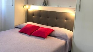 Una cama con cabecera capitonada, un almohadón rojo y una repisa encima.