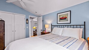 A bedroom with a bed, a ceiling fan, a nightstand with a lamp, a framed picture on the wall, and a door leading to another room.