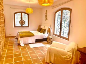 Ein Schlafzimmer mit einem Bett, einem Nachttisch, einem Sessel und zwei Fenstern mit schmiedeeisernen Gitterrosten.