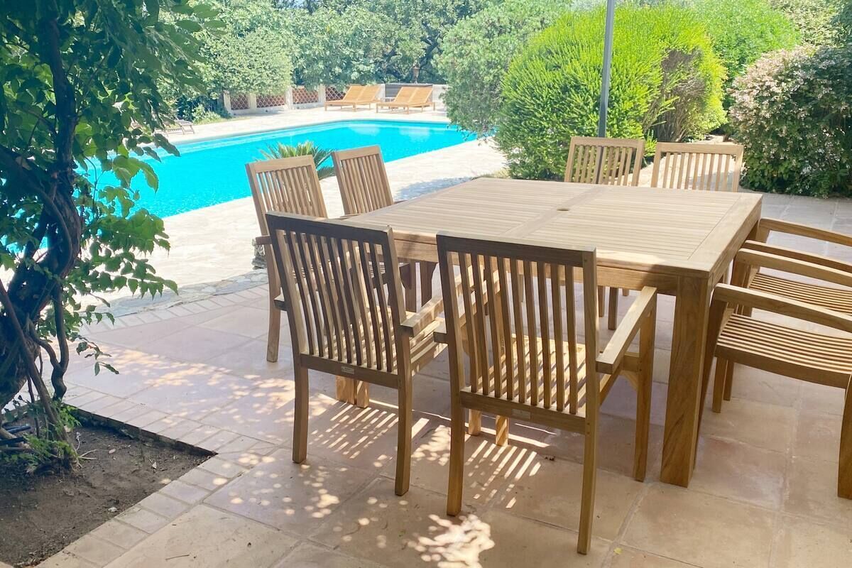 Une table et des chaises en bois sont disposées sur une terrasse, avec une piscine en arrière-plan.
