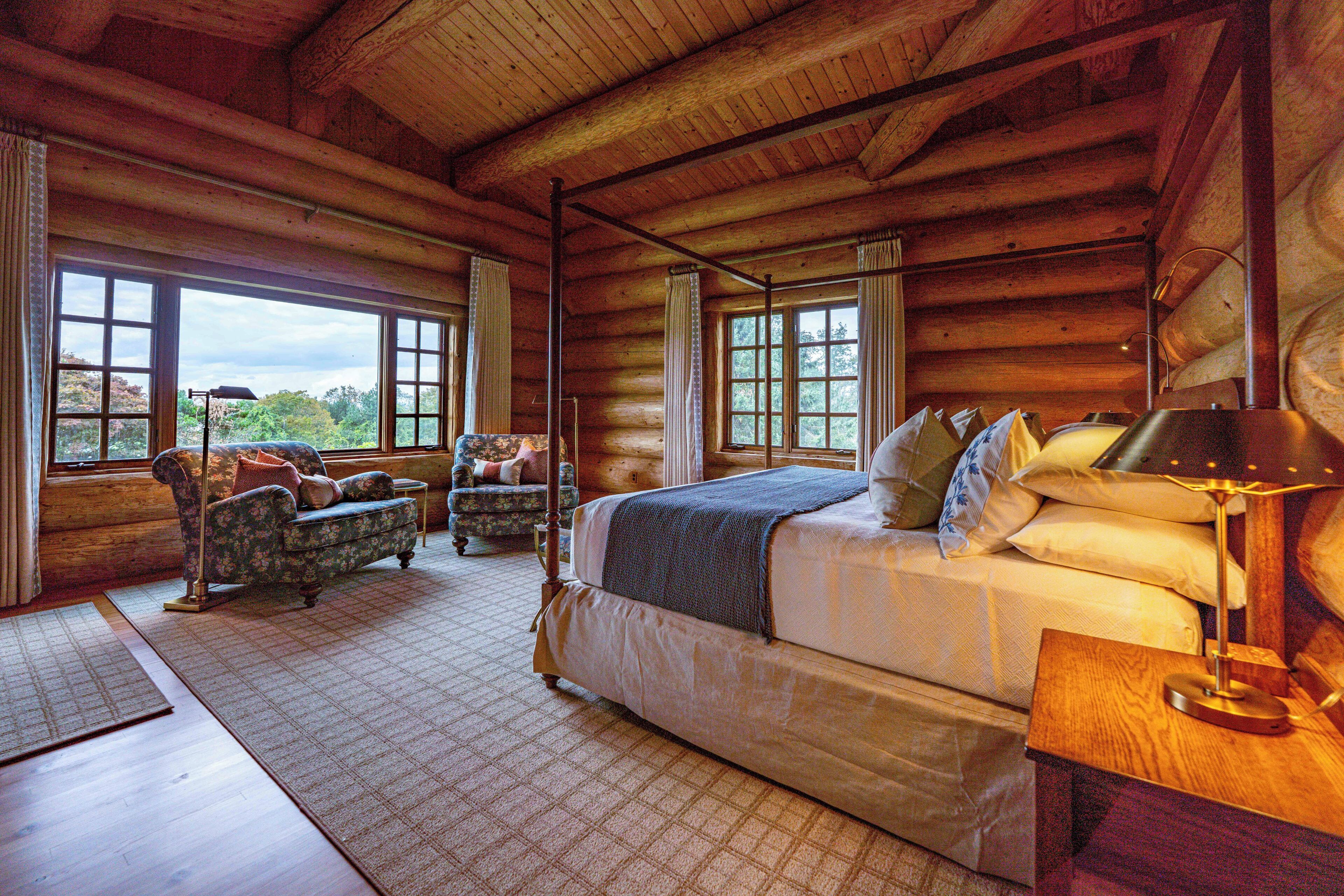 Une chambre située dans un chalet en rondins, avec un grand lit, deux fauteuils et une vue sur l’extérieur grâce à de grandes fenêtres.