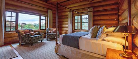 Une chambre située dans un chalet en rondins, avec un grand lit, deux fauteuils et une vue sur l’extérieur grâce à de grandes fenêtres.