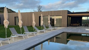 Un área de piscina moderna al aire libre con sillones de descanso y sombrillas, una terraza de madera y un edificio de diseño contemporáneo.