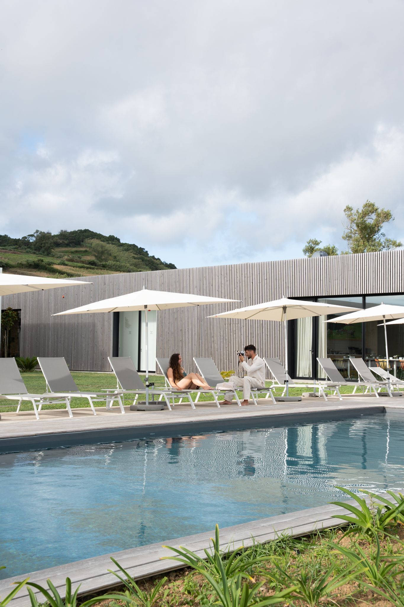 A pool area with lounge chairs and umbrellas, a modern building in the background, and a clear sky.