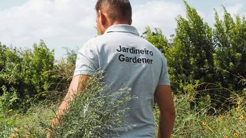 Eine Person mit T-Shirt, auf dessen Rückseite 'Jardineiro Gardener' steht, steht in einer natürlichen Umgebung mit Grünpflanzen und Blumen.