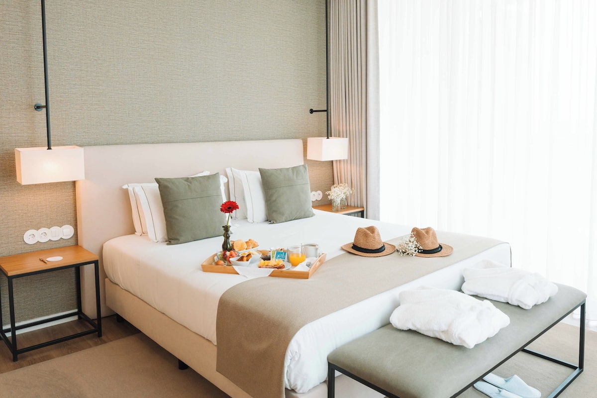 Habitación de hotel con una cama grande, dos mesitas de noche, una lámpara de noche, una bandeja con alimentos para el desayuno, un sombrero y toallas.