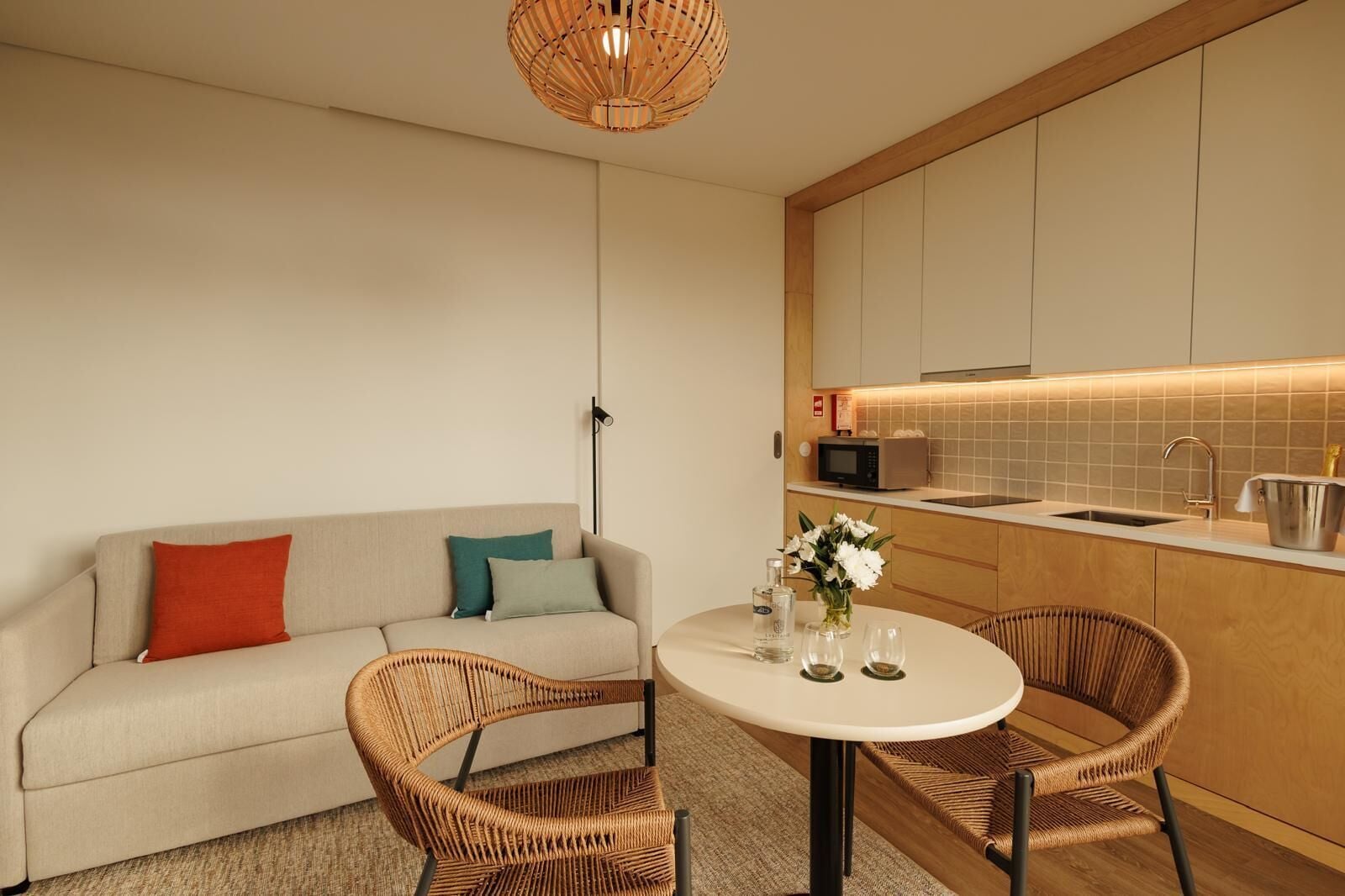 A modern, compact living space with a sofa, two wicker chairs, a small round table, a kitchen area with a microwave, and a hanging light fixture.