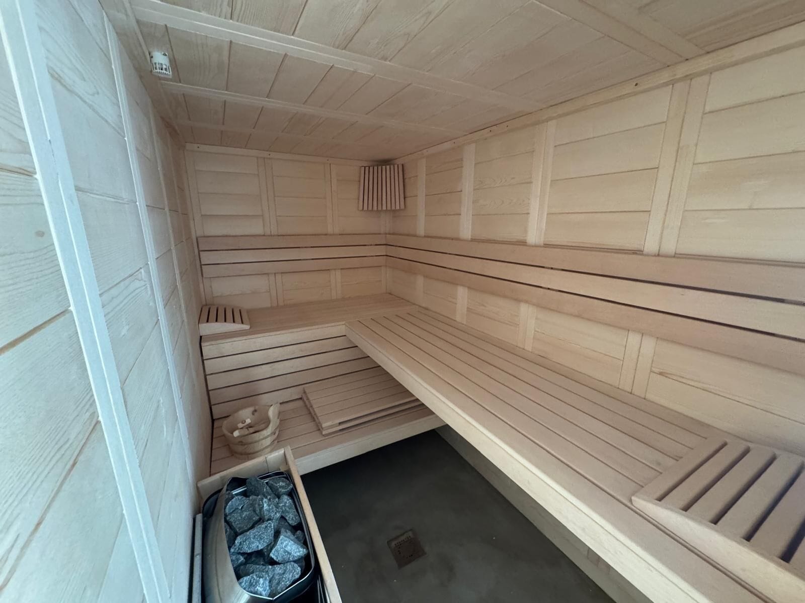 A sauna with wooden benches and a bench for sitting. There is a small trash bin and a heater visible.